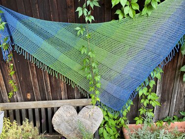 Blau-grünes gestricktes Dreieckstuch mit Lochmuster und Fransen, an einem Holzzaun aufgehängt.