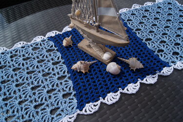 Gehäkelter Tischläufer in Blau- und Weißtönen mit Muscheln und Holzsegelschiff auf dunklem Tisch