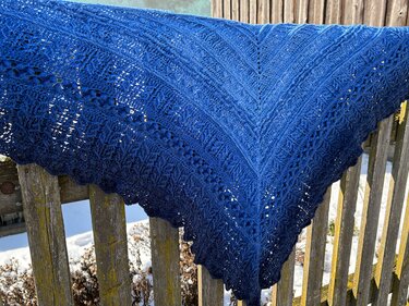 Blauer gehäkelter Dreiecksschal mit Spitzen- und Noppenmuster auf einem Holzzaun, Schnee im Hintergrund