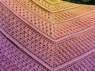 Crocheted triangle shawl with a gradient from yellow to purple, showing lacy openwork stitches and textured stitch bands.
