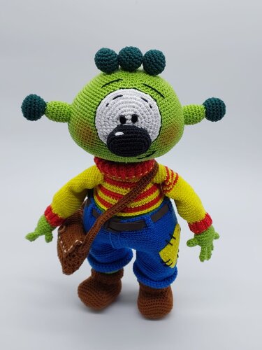 Gehäkeltes grünes Amigurumi-Monster mit großer Augenpartie, Fühlern, gestreiftem Shirt, blauer Hose, Stiefeln und Umhängetasche