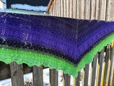 Gehäkeltes Dreieckstuch in Violett und Grün mit Noppenmuster und Muschelrand auf einem Holzzaun