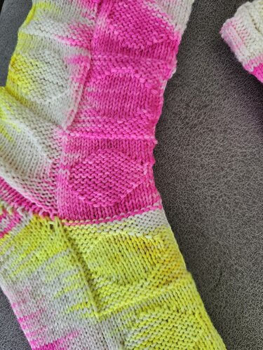 Handgestrickte Socke in Neonpink, Neongelb und Weiß mit geometrischen, erhabenen Quadraten auf grauem Untergrund.