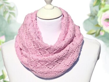 Rosa gestrickter Loop-Schal mit Lochmuster auf einer Schaufensterpuppe vor hellem, floralem Hintergrund.