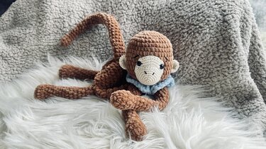 Handgehäkelter brauner Amigurumi-Affe mit beigem Gesicht und hellblauem Kragen auf weißem Kunstfell
