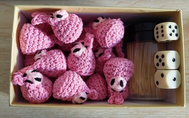 Kleine rosafarbene gehäkelte Amigurumi-Figuren in einer Schachtel neben drei Würfeln