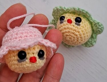 Hand hält kleines gelbes gehäkeltes Küken mit rosa Mütze; weiteres Küken mit grüner Mütze im Hintergrund.