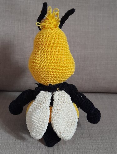 Rückansicht einer gehäkelten gelb-schwarzen Amigurumi-Biene mit weißen Flügeln auf hellem Stoffhintergrund.