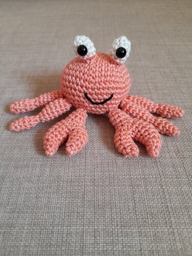 Gehäkelte korallenrosa Krabbe mit acht Beinen, weißen Augenstielen und gesticktem schwarzem Lächeln auf beigem Stoff