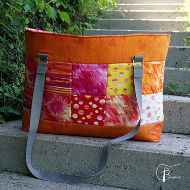 Bunte Patchwork-Tasche mit orangefarbener Basis und grauen Trageriemen auf Steinstufen.