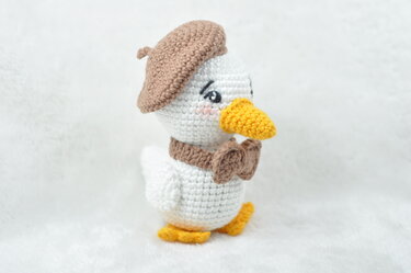 White crocheted duck amigurumi wearing a brown beret and bow tie, with a yellow beak and feet on a white background.