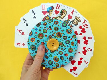 Hand hält runden blauen Stoff-Kartenhalter mit bunten Kreisen und gelbem Knopf; dahinter gefächerte Spielkarten.