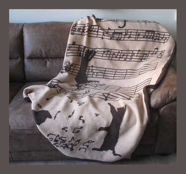 Beige gehäkelte Decke mit schwarzen Musiknoten und Katzen auf einem braunen Sofa