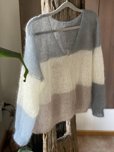 Gestreifter Strickpullover mit V-Ausschnitt in Hellblau, Weiß und Taupe, an einem Holzstamm hängend.