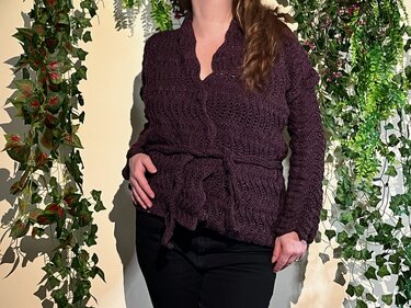 Person wearing a dark plum crocheted jacket with tie belt, long sleeves and notched neckline against a leafy background