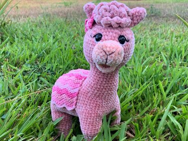 Pink crocheted alpaca-style amigurumi with scalloped saddle standing on green grass