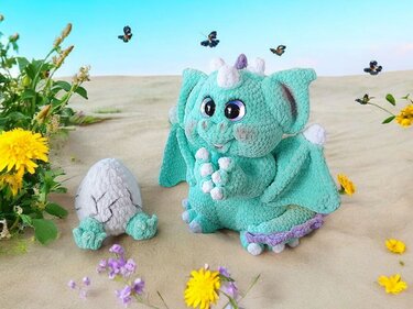 Mint green crochet dragon plush beside a cracked crochet egg on sand with flowers and butterflies.