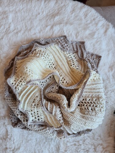 Gehäkelter Drachenschwanz-Schal in Creme und Taupe mit Rüschenrand, zusammengerollt auf hellem Felluntergrund