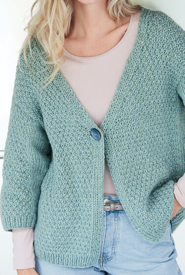 Dame trägt salbeigrüne, grob gestrickte Kurzjacke mit V‑Ausschnitt und großem Knopf, kombiniert mit hellem Shirt und Jeans