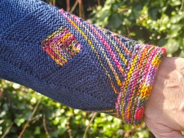 Nahaufnahme eines dunkelblauen gestrickten Ärmels mit bunt gestreiftem Bündchen und kleinem quadratischen Detail