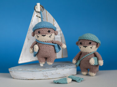 Two small crocheted otter amigurumi keychains with teal hats and tiny fish accessories against a blue background and wooden sailboat prop