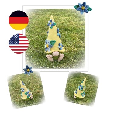 Crocheted gnome with a tall yellow hat decorated with blue flowers, green body, beige nose, and feet on grass.