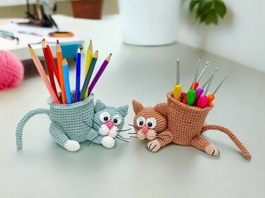 Zwei gehäkelte Katzen-Utensilos (hellblau und braun) mit Buntstiften und Häkelnadeln auf einem Schreibtisch.