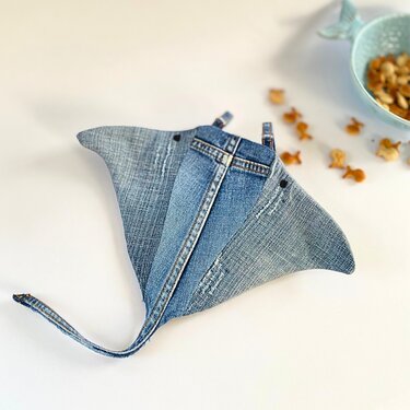 Rochenförmiger Topflappen aus Jeansstoff mit Aufhängeschlaufe auf hellem Untergrund, umgeben von Fischcrackern