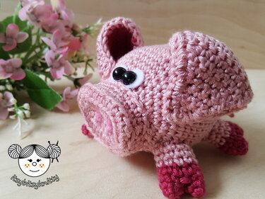 Rosa gehäkeltes Schwein mit schwarzen Augen und dunklen Hufen auf hellem Holz, mit rosa Blüten im Hintergrund.