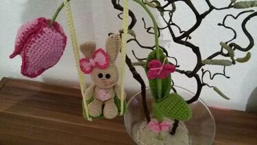 Gehäkelter beiger Amigurumi-Hase auf Schaukel mit rosa Tulpe und Schleifen an Zweig