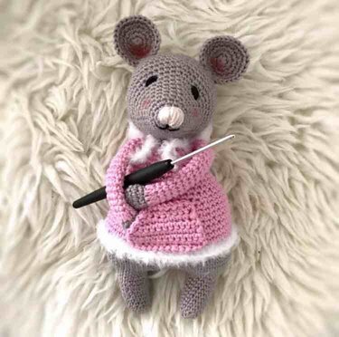 Grau gehäkelte Amigurumi‑Maus mit rosa Mantel und Häkelnadel auf flauschigem Teppich