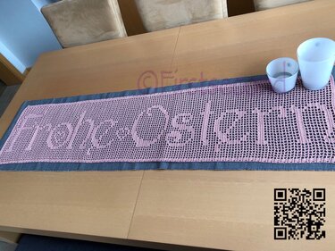 Rosa Filet-Häkel-Tischläufer mit Schriftzug ‚Frohe Ostern‘ auf hellem Holztisch, rechts zwei weiße Deko-Becher