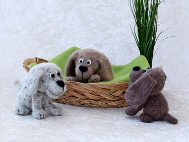 Drei gehäkelte Amigurumi-Welpen: zwei vor einem Weidenkorb, einer darin auf grünem Tuch, Topfpflanze im Hintergrund