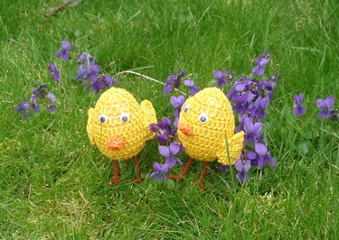 Zwei handgehäkelte gelbe Küken mit orangen Schnäbeln zwischen violetten Veilchen auf grünem Gras