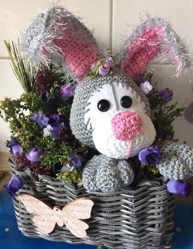 Gehäkelter grauer Amigurumi‑Hase mit rosa Nase und flauschigen Ohren in grauem Korb mit violetten Blumen