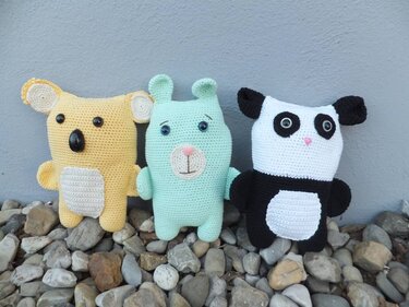 Drei gehäkelte Amigurumi: gelber Koala, mintgrüner Bär und schwarz-weißer Panda auf Kieselsteinen vor einer grauen Wand.