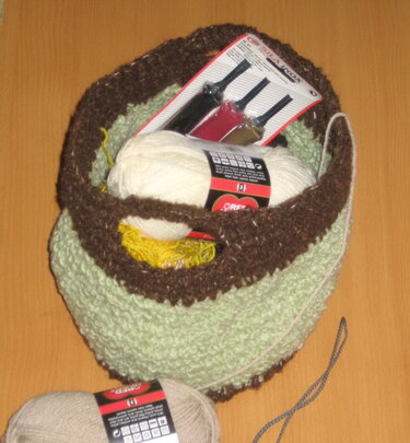 Light green and brown crochet basket holding white yarn and a pack of three crochet hooks, with a beige skein nearby.