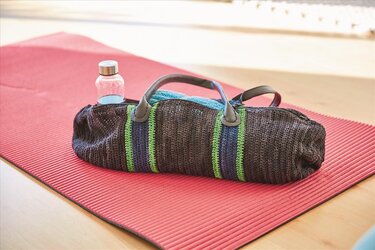 Gehäkelte dunkelgraue Sporttasche mit grünen und blauen Streifen auf roter Yogamatte, Trinkflasche im Fach
