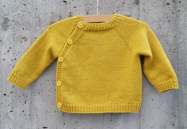 Helle gestrickte Babyjacke in Gelb mit seitlicher Knopfleiste und Raglanärmeln an einem Holzbügel vor einer grauen Wand.