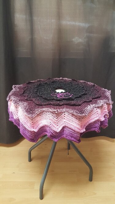 Runde gehäkelte Decke mit Farbverlauf in Schwarz, Lila und Pink auf dreibeinigem Hocker