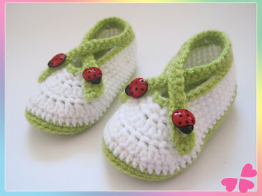Gehäkelte weiße Babyschuhe mit hellgrünen Riemchen und roten Marienkäfer-Knöpfen auf weißem Hintergrund.