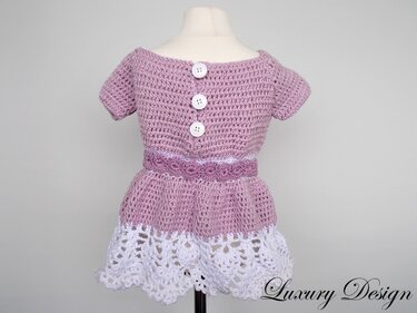 Gehäkeltes Kinderkleid in Lila mit weißer Borte und drei weißen Knöpfen am Rücken auf einer Schaufensterpuppe.