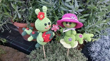 Zwei handgehäkelte grüne Amigurumi-Gartenstecker: Raupe mit Erdbeere und Figur mit pinkem Hut zwischen Pflanzen