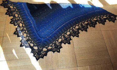 Dreieckiges gehäkeltes Tuch in Blau bis Schwarz mit schwarzer Spitzenborte auf Parkettboden, teils im Sonnenlicht