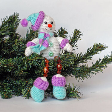 Crocheted snowman amigurumi wearing a purple and turquoise striped hat and scarf on pine branches.