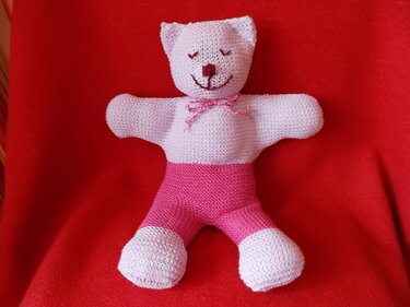 Gestrickter Teddybär in Lila und Rosa mit gesticktem Gesicht und kleiner Schleife auf rotem Hintergrund.