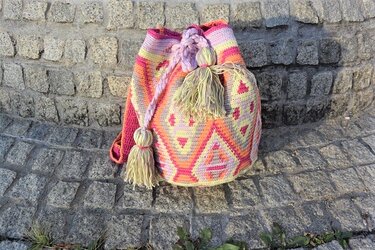 Gehäkelte Umhängetasche in Pink, Orange und Gelb mit geometrischem Muster und Quasten auf einer Steinmauer.