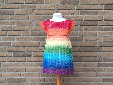 Gehäkeltes Kinderkleid in Regenbogenfarben auf einer Schaufensterpuppe vor einer Ziegelwand