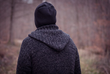 Rückansicht: dunkelgraue, grob gehäkelte Kapuzenjacke mit schwarzer Beanie vor unscharfem Herbstwald.