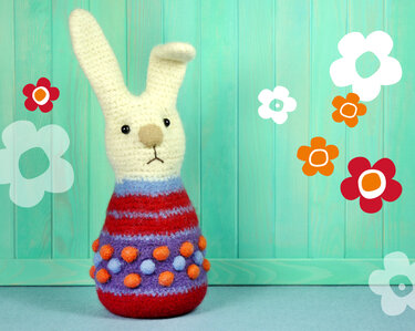 Gehäkelter Hase mit cremefarbenem Kopf und bunt gestreiftem, gefilztem Körper vor türkisfarbener Holzwand mit Blumenmotiven